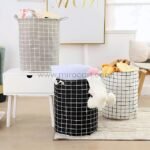 Set of three cylindrical laundry bags with grid pattern in grey, black, and white, featuring carry handles in a cozy room setting.