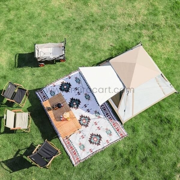 Aerial view of a camping setup with a beige tent, patterned rug, wooden table, folding chairs, and a collapsible wagon.