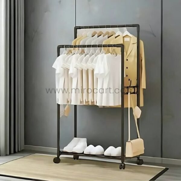 Black space-saving clothes rack holding neatly arranged shirts, with shoes below and a bag on the side, set against gray walls and sheer curtains.