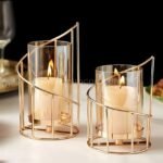 Golden pillar candle holder with spiral metal frame and glass hurricane cover glowing softly on marble surface.