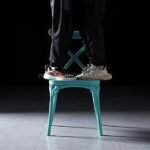 Two adults standing on one turquoise cross-back chair.