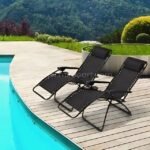 Two sleek black zero-gravity loungers rest on a sun-warmed wooden deck, framing a glistening pool with lush greenery and endless blue skies.