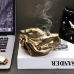 A bronze creative hand ashtray with a cigarette resting on it, placed on a minimalist desk.