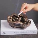 A hand placing a lit cigarette into a bronze creative hand ashtray.