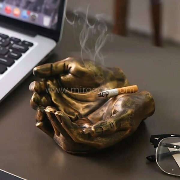 A bronze creative hand ashtray with a cigarette resting on top, placed casually on a table.