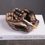 A bronze creative hand ashtray with a cigarette resting on top, placed on an open book.