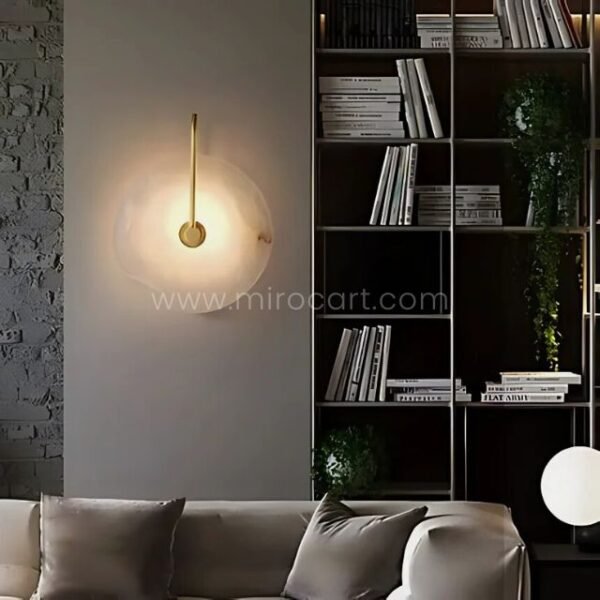 Gold-accented alabaster wall light near a bookshelf and greenery, casting warm light in a cozy nook.