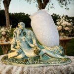 Sculpted lovers figurine with opaline glass vase placed on a stone garden table surrounded by blooming flowers and greenery.