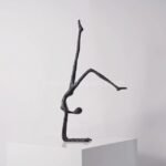 Sleek black metal sculpture of a human figure performing a handstand, displayed on a white pedestal against a clean, neutral background.