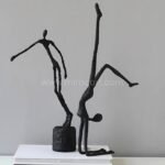 Two black metal sculptures of human figures in dynamic poses, one standing and one performing a handstand, displayed together on a white pedestal.