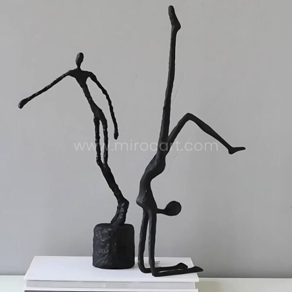 Two black metal sculptures of human figures in dynamic poses, one standing and one performing a handstand, displayed together on a white pedestal.
