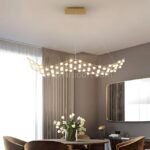 Flowing gold chandelier with organic curves glowing above a sleek dining table, framed by neutral drapes and a modern mirror.