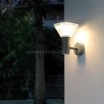 Gray modern outdoor wall light mounted on a white garden wall surrounded by plants.