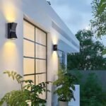 Modern up and down wall light mounted on a modern home exterior with potted plants and large windows.