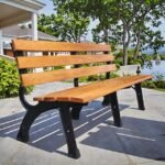 A classic wooden park bench with a black metal frame sits on a rustic stone patio at the edge of a calm lake, surrounded by lush green trees.