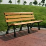 Classic wooden park bench with a light brown solid wood seat and black metal frame, situated on a red composite deck overlooking a green lawn.