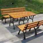 Two classic wooden park benches with a matching wood-and-metal table create an outdoor seating area on a paved patio next to a manicured lawn.