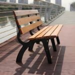 A classic wooden park bench with a black metal frame sits on a scenic red composite riverwalk deck next to a railing, overlooking the water.